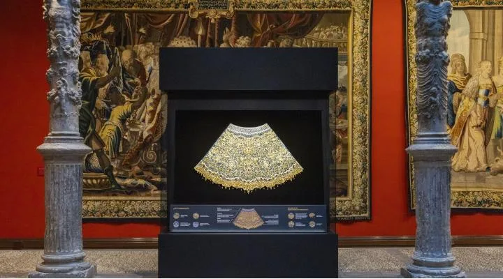 Gold, ornate necklace on a black display case against a red museum wall with tapestries in the background