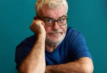 Older man with gray hair and beard wearing glasses rests his head on his hand against a teal background