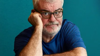 Older man with gray hair and beard wearing glasses rests his head on his hand against a teal background