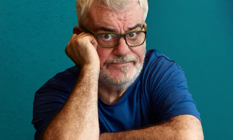 Older man with gray hair and beard wearing glasses rests his head on his hand against a teal background