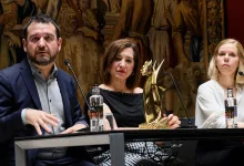 Three panelists at a conference table; center woman holds a gold trophy while the man on the left speaks into a microphone and the woman on the right looks ahead, with a decorative mural behind them.