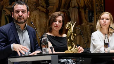 Three panelists at a conference table; center woman holds a gold trophy while the man on the left speaks into a microphone and the woman on the right looks ahead, with a decorative mural behind them.