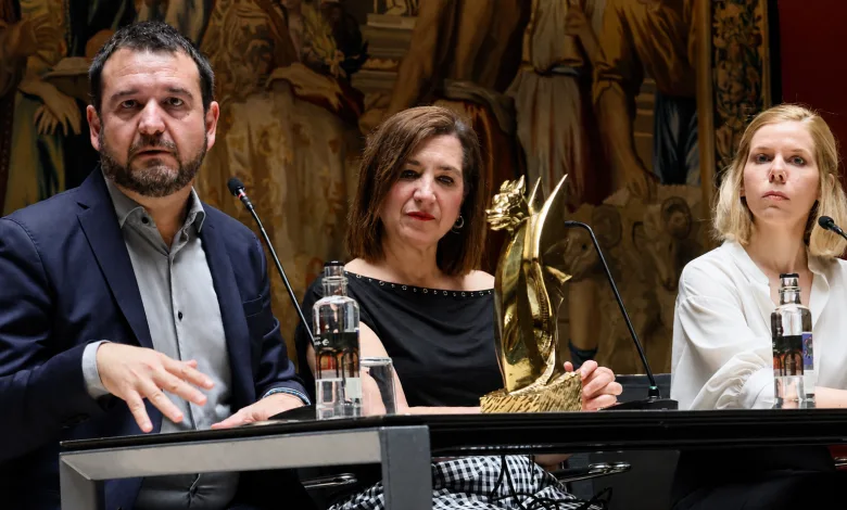 Three panelists at a conference table; center woman holds a gold trophy while the man on the left speaks into a microphone and the woman on the right looks ahead, with a decorative mural behind them.
