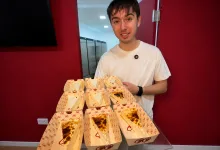 Young Man in a White T shirt Stands Behind a Table Displaying Multiple Opened Pizza Boxes Arranged Like Wedges on a Counter in a Red walled Room