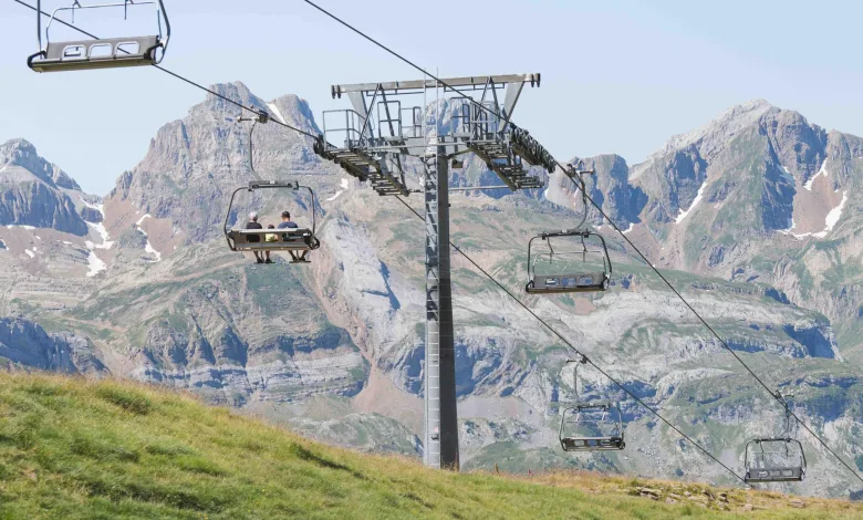 Chairlift Carrying Three Riders over a Green Hillside with Rugged Alpine Mountains in the Background Astún Abre El Verano Con El Telesilla Los Lagos Zaragozala   Qué Hacer En Zaragoza