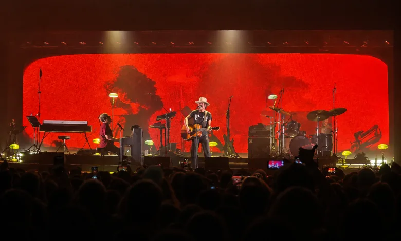 Guitarist in a Hat Center Stage Playing an Acoustic Guitar During a Live Concert with a Red Abstract Backdrop and Band Members Nearby Audience in the Foreground Iván Ferreiro Repasa Sus Grandes Clásicos En La Multiusos Zaragozala   Qué Hacer En Zaragoza