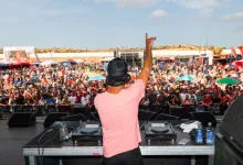 Dj on Stage with Pink Shirt Raises Right Arm to Cheering Crowd at a Large Outdoor Festival Mixing Gear in Front of Him Zaragozala | Agenda Cultural Ocio Y Planes En Zaragoza Zaragozala   Qué Hacer En Zaragoza
