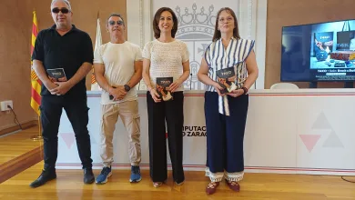 Four Adults Stand in a Row Indoors at a Diputación De Zaragoza Setting Each Holding a Brochure and Smiling at the Camera with a Spanish Flag and a Screen in the Background Tako Celebra 40 Años Con Libro Y Homenaje a Labordeta Zaragozala   Qué Hacer En Zaragoza
