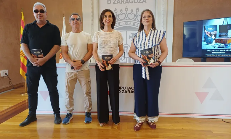 Four Adults Stand in a Row Indoors at a Diputación De Zaragoza Setting Each Holding a Brochure and Smiling at the Camera with a Spanish Flag and a Screen in the Background Tako Celebra 40 Años Con Libro Y Homenaje a Labordeta Zaragozala Qué Hacer En Zaragoza
