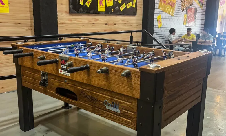 Indoor Foosball Table with a Wooden Finish and Black Legs Players Are on the Rods in a Space with Yellow Posters on the Wall at the Back Comer Jugar Y Celebrar Así Convierten Dos Locales De Zaragoza El Mundial En Una Competición De Barrio Zaragozala   Qué Hacer En Zaragoza