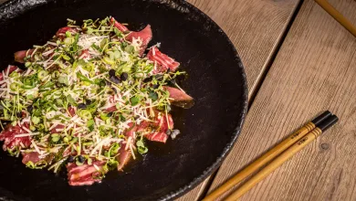 Carpaccio style Beef on a Black Plate Topped with Microgreens and Grated Cheese Set on a Wooden Table with Chopsticks Nearby La Cocina Japonesa En Zaragoza Suma Nuevos Platos Zaragozala   Qué Hacer En Zaragoza