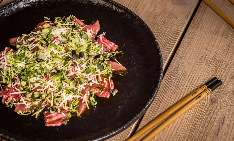 Carpaccio style Beef on a Black Plate Topped with Microgreens and Grated Cheese Set on a Wooden Table with Chopsticks Nearby La Cocina Japonesa En Zaragoza Suma Nuevos Platos Zaragozala   Qué Hacer En Zaragoza