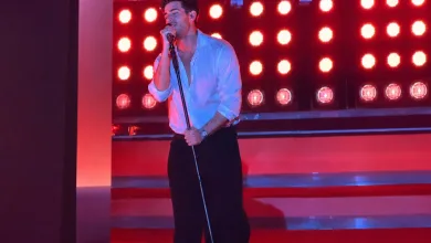 Male singer on stage, wearing a white shirt and black pants, singing into a mic under red stage lights.