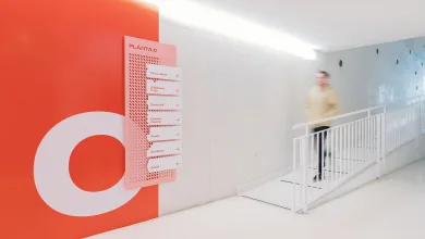 Pink Directional Signboard with Multiple Arrows Mounted on an Orange Wall in a Bright Modern Hallway a Blurred Person Walks Past a White Railing La Señalética De Zaragoza Joven Gana Un Premio Europeo Zaragozala Qué Hacer En Zaragoza