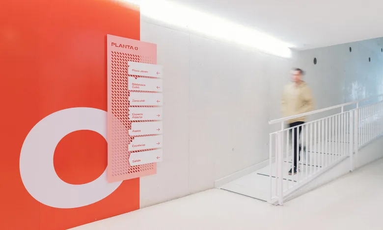 Pink Directional Signboard with Multiple Arrows Mounted on an Orange Wall in a Bright Modern Hallway a Blurred Person Walks Past a White Railing La Señalética De Zaragoza Joven Gana Un Premio Europeo Zaragozala Qué Hacer En Zaragoza