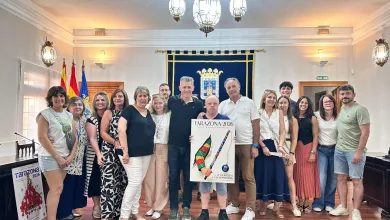 Group of about 18 adults posing for a photo in a formal room, with a blue crest banner and flags in the background, holding a Tarazona 2026 poster.