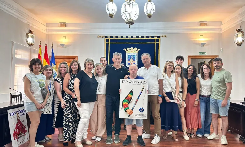Group of About 18 Adults Posing for a Photo in a Formal Room with a Blue Crest Banner and Flags in the Background Holding a Tarazona 2026 Poster rompiendo Moldes El Cartel Que Anunciará Las Fiestas De Tarazona 2026 Zaragozala Qué Hacer En Zaragoza