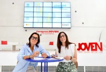 Two Women Sit at a Blue Round Table Signing Papers in a Bright Modern Space with a Large Digital Screen Behind Them and zaragoza Joven Signage in Red Zaragozala | Agenda Cultural Ocio Y Planes En Zaragoza Zaragozala Qué Hacer En Zaragoza