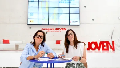 Two Women Sit at a Blue Round Table Signing Papers in a Bright Modern Space with a Large Digital Screen Behind Them and zaragoza Joven Signage in Red Loading Verano De Zaragoza Joven Ofrece 11 Camps De Verano Para Jóvenes Zaragozala Qué Hacer En Zaragoza