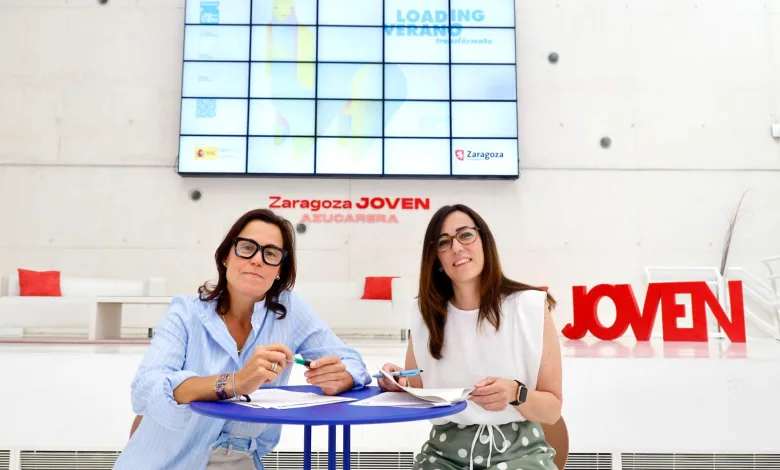 Two Women Sit at a Blue Round Table Signing Papers in a Bright Modern Space with a Large Digital Screen Behind Them and zaragoza Joven Signage in Red Zaragozala | Agenda Cultural Ocio Y Planes En Zaragoza   Página 1404 Zaragozala   Qué Hacer En Zaragoza