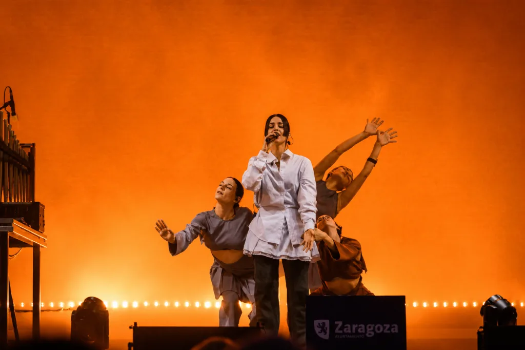 Female Singer in a White Shirt Performs Center Stage with a Microphone Surrounded by Three Expressive Dancers Against an Bright Orange Backdrop Stage Lighting and Equipment Visible on the Sides María Arnal Y Nortec Ponen El Broche Final a Música Al Raso Zaragozala Qué Hacer En Zaragoza