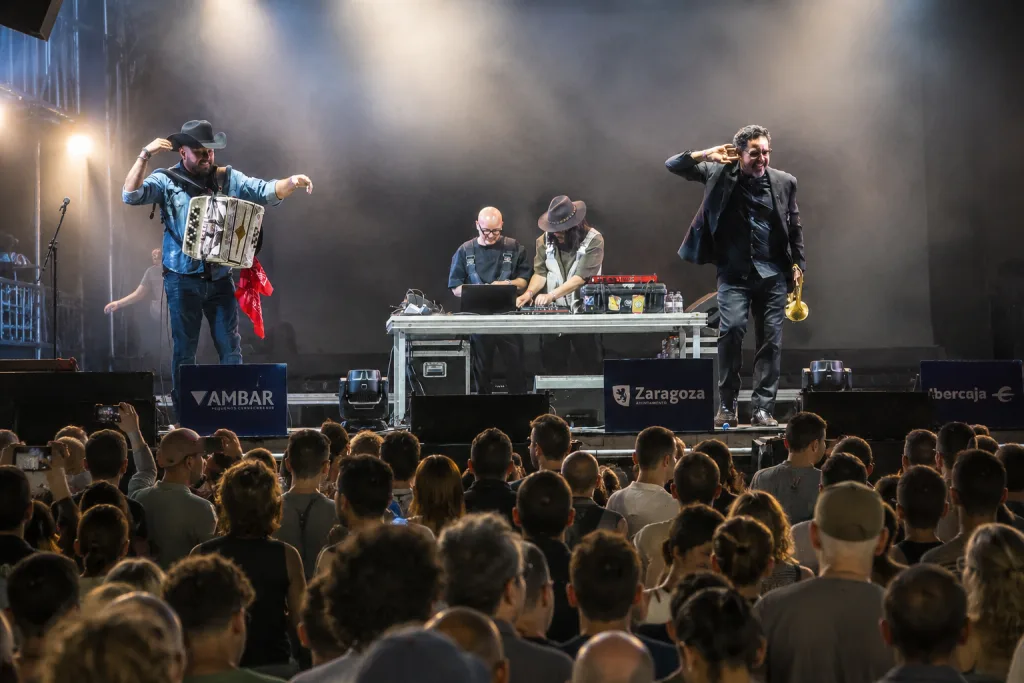 Band Performing on a Stage with a Man Playing an Accordion a Dj at a Mixing Table and a Horn Player in a Black Suit Crowd Watching in Front of the Stage María Arnal Y Nortec Ponen El Broche Final a Música Al Raso Zaragozala Qué Hacer En Zaragoza