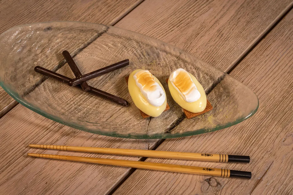 Two Yellow Oval Pastries with Cream and Toasted Tops on a Glass Plate with Chocolate Sticks and Chopsticks Nearby on a Wooden Table La Cocina Japonesa En Zaragoza Suma Nuevos Platos Zaragozala   Qué Hacer En Zaragoza
