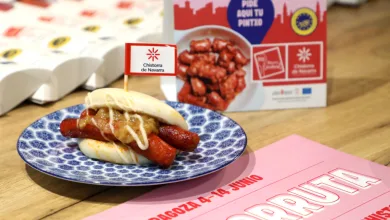 Plate with a Bun Filled with Chistorra Sausages and Sauce Topped with a Small Flag Reading chistorra De Navarra Set on a Blue Patterned Plate at a Food Event Banquet Area La Ruta De La Chistorra De Navarra Llega a Zaragoza Zaragozala   Qué Hacer En Zaragoza