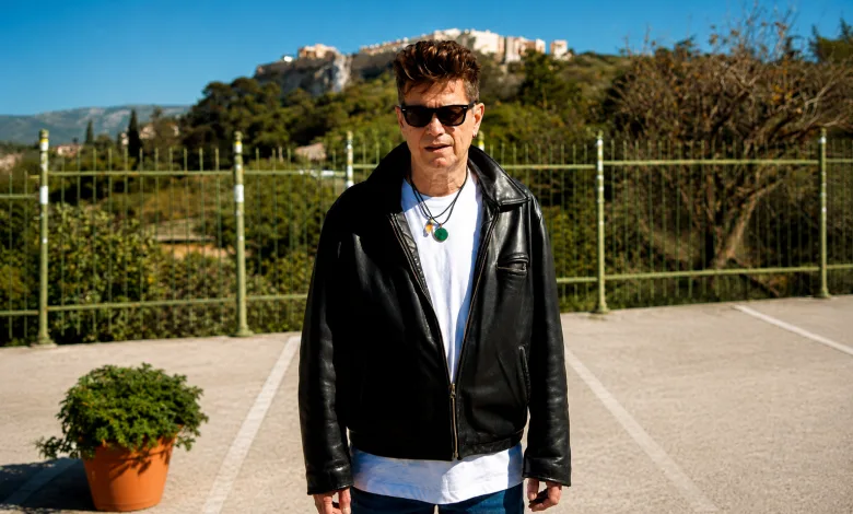 Man in a black leather jacket and white shirt stands in a parking lot with a green fence and hillside behind him.