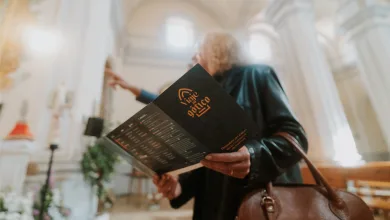 Person in Dark Clothing Holds a Black Brochure with an Orange Logo in a Bright Arched Hallway a Brown Bag is in View Viaje Al Gótico 2026 Vuelve a Campo De Daroca Zaragozala   Qué Hacer En Zaragoza