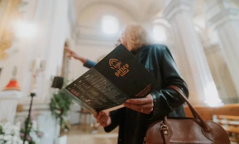 Person in Dark Clothing Holds a Black Brochure with an Orange Logo in a Bright Arched Hallway a Brown Bag is in View Viaje Al Gótico 2026 Vuelve a Campo De Daroca Zaragozala   Qué Hacer En Zaragoza