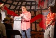 Man in Coral Shirt Speaks at a Transparent Podium with Two Microphones Flanked by an Older Man in a Gray Suit and a Woman in a Red Patterned Blouse Zaragoza Ayuntamiento Sign on the Lectern Zaragozala | Agenda Cultural Ocio Y Planes En Zaragoza Zaragozala   Qué Hacer En Zaragoza