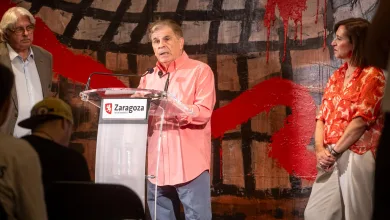 Man in Coral Shirt Speaks at a Transparent Podium with Two Microphones Flanked by an Older Man in a Gray Suit and a Woman in a Red Patterned Blouse Zaragoza Ayuntamiento Sign on the Lectern Zaragozala | Agenda Cultural Ocio Y Planes En Zaragoza Zaragozala   Qué Hacer En Zaragoza