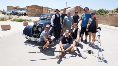 Film crew posing beside a classic black car on a sunny street, with gear and a tripod nearby.