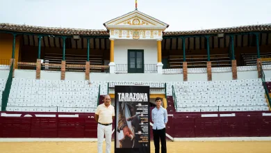 Two Men Posing Beside a Tall Poster for the Tarazona Bullfighting Festival in Front of Stadium Seating and a Decorative Building with a Flag on Top Feria Taurina De Tarazona 2026 Roca Rey Lidera El Cartel Zaragozala Qué Hacer En Zaragoza