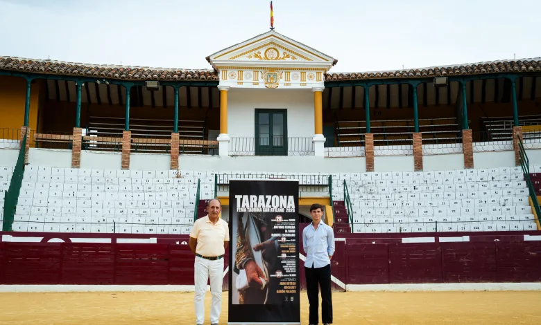 Two Men Posing Beside a Tall Poster for the Tarazona Bullfighting Festival in Front of Stadium Seating and a Decorative Building with a Flag on Top Feria Taurina De Tarazona 2026 Roca Rey Lidera El Cartel Zaragozala Qué Hacer En Zaragoza