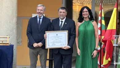 Man in a Dark Suit Holding a Framed Certificate Flanked by Two Colleagues at an Awards Ceremony with Colorful Flags in the Background Ernesto Escar Parrilla De Oro De Huesca 2026 Zaragozala   Qué Hacer En Zaragoza