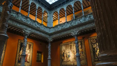 Historic Gallery Interior with Orange Walls Carved Arches and Grey Stone Columns Surrounding Framed Tapestries Under a Skylight La Carta Astral Oculta En El Patio De La Infanta Zaragozala   Qué Hacer En Zaragoza