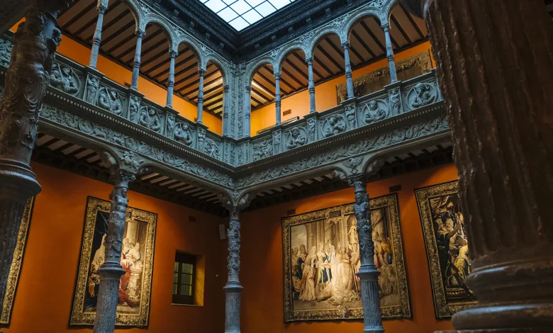 Historic Gallery Interior with Orange Walls Carved Arches and Grey Stone Columns Surrounding Framed Tapestries Under a Skylight La Carta Astral Oculta En El Patio De La Infanta Zaragozala   Qué Hacer En Zaragoza