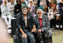 Two Smiling Older Performers in Costumes Sit on a Wooden Chest at an Outdoor Event Holding Hands As a Crowd Applauds Behind Them Zaragozala | Agenda Cultural Ocio Y Planes En Zaragoza Zaragozala Qué Hacer En Zaragoza