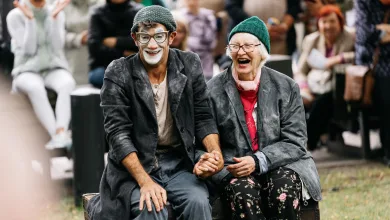 Two Smiling Older Performers in Costumes Sit on a Wooden Chest at an Outdoor Event Holding Hands As a Crowd Applauds Behind Them Festival Ambulantes Noches De Circo En Jardín De Las Artes Zaragozala Qué Hacer En Zaragoza