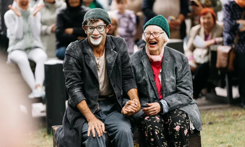Two Smiling Older Performers in Costumes Sit on a Wooden Chest at an Outdoor Event Holding Hands As a Crowd Applauds Behind Them Festival Ambulantes Noches De Circo En Jardín De Las Artes Zaragozala Qué Hacer En Zaragoza