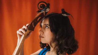 Portrait of a Woman with Two Small Birds Perched on Her Head Holding a Large Wooden String Instrument Against an Orange Backdrop Pilar Almalé Y Miguel Tena Abren El Ciclo en Torno Al Agua Zaragozala Qué Hacer En Zaragoza