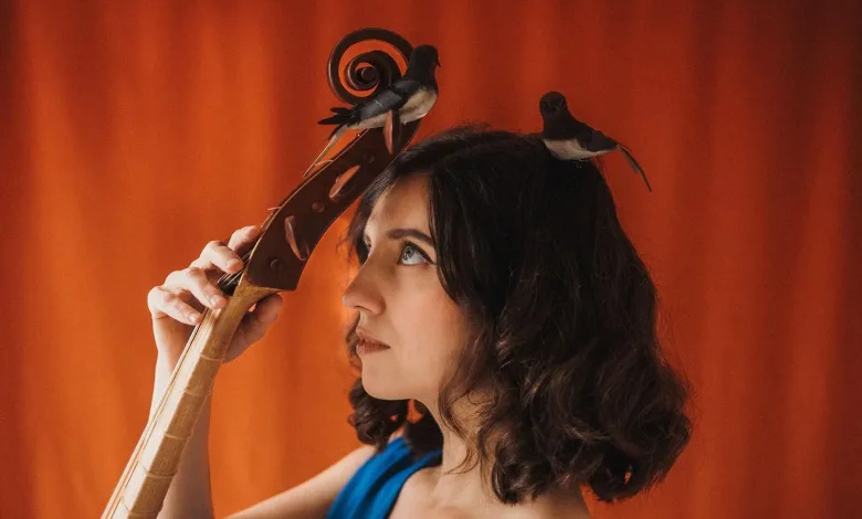 Portrait of a Woman with Two Small Birds Perched on Her Head Holding a Large Wooden String Instrument Against an Orange Backdrop Pilar Almalé Y Miguel Tena Abren El Ciclo en Torno Al Agua Zaragozala Qué Hacer En Zaragoza
