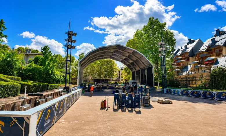 Open air Stage with a Curved Metal Canopy Tall Lighting Rigs and Stacks of Blue Chairs Under a Bright Blue Sky El Fiestón Del Verano Jaca 2026 Queda Cancelado Zaragozala Qué Hacer En Zaragoza