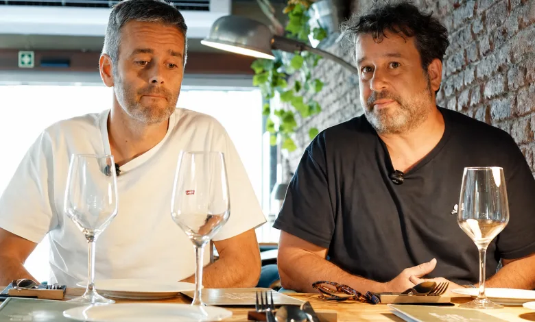 Two Men Sit at a Brick walled Restaurant Table with Empty Wine Glasses in Front of Them Appearing to Chat Seriously One Wears a White Shirt the Other a Black Shirt Reservas Fantasma En Zaragoza El Caso Viral De La Miguería Zaragozala   Qué Hacer En Zaragoza