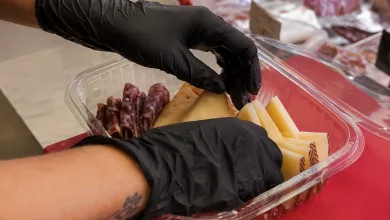 Gloved hands arrange cheese and salami slices in a clear plastic container on a red surface.