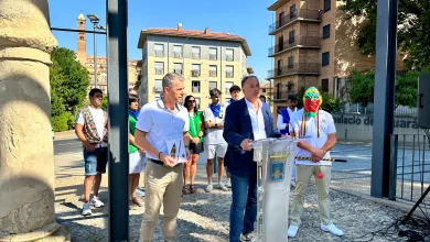 Man in a Blazer Speaks at a Clear Podium During an Outdoor Ceremony with a Colorful Masked Performer and Onlookers in a Sunny Urban Square Behind Him Tarazona Presenta Las Fiestas 2026 Y Su Cipotegato Zaragozala Qué Hacer En Zaragoza