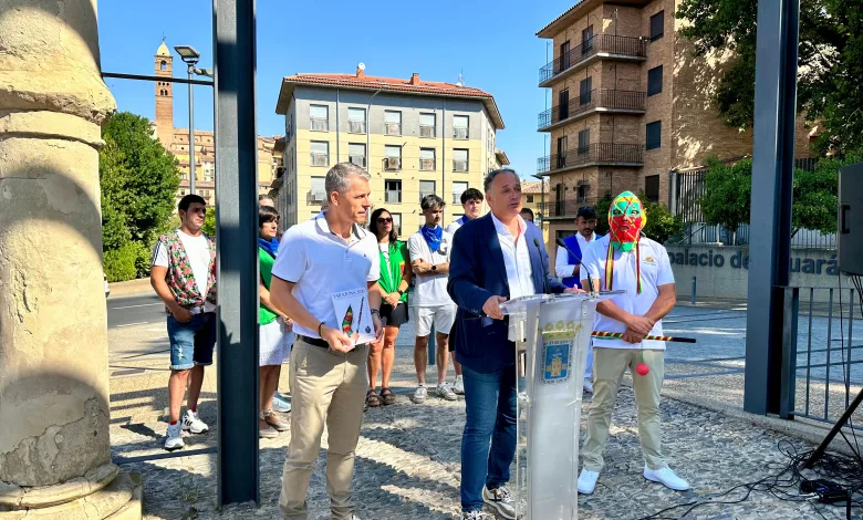 Man in a Blazer Speaks at a Clear Podium During an Outdoor Ceremony with a Colorful Masked Performer and Onlookers in a Sunny Urban Square Behind Him Tarazona Presenta Las Fiestas 2026 Y Su Cipotegato Zaragozala Qué Hacer En Zaragoza